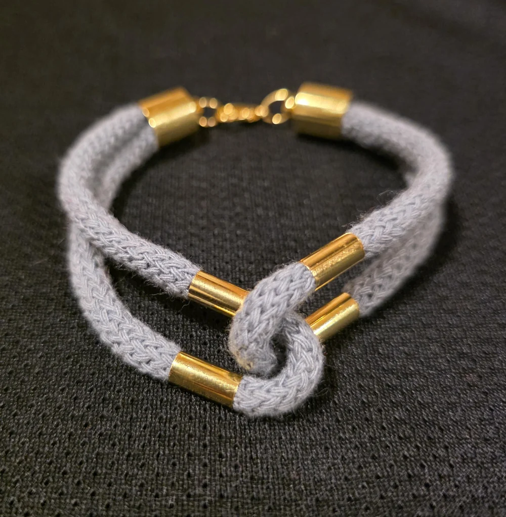 A pale grey knotted cord bracelet with gold-coloured details.