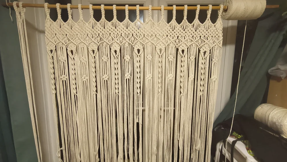 A cream macramé curtain with long fringe hanging from a wooden rod.