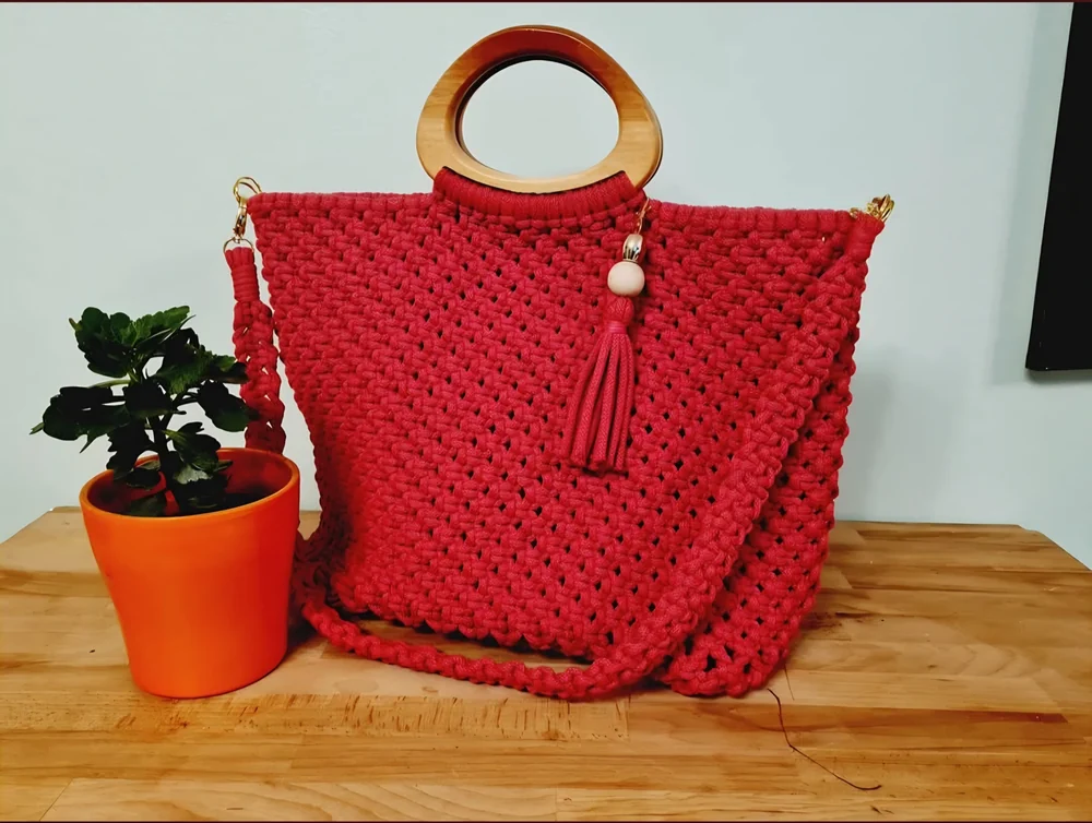 A red knotted macramé bag with round wooden handles beside a plant.